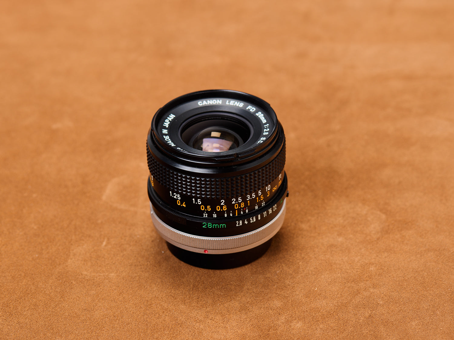 Canon 28mm f/2.8 FD, black barrel stretched on tan suede. Chrome aperture ring flashes silver. Distance scales whisper alibis. Narrow your eyes. Touch it.