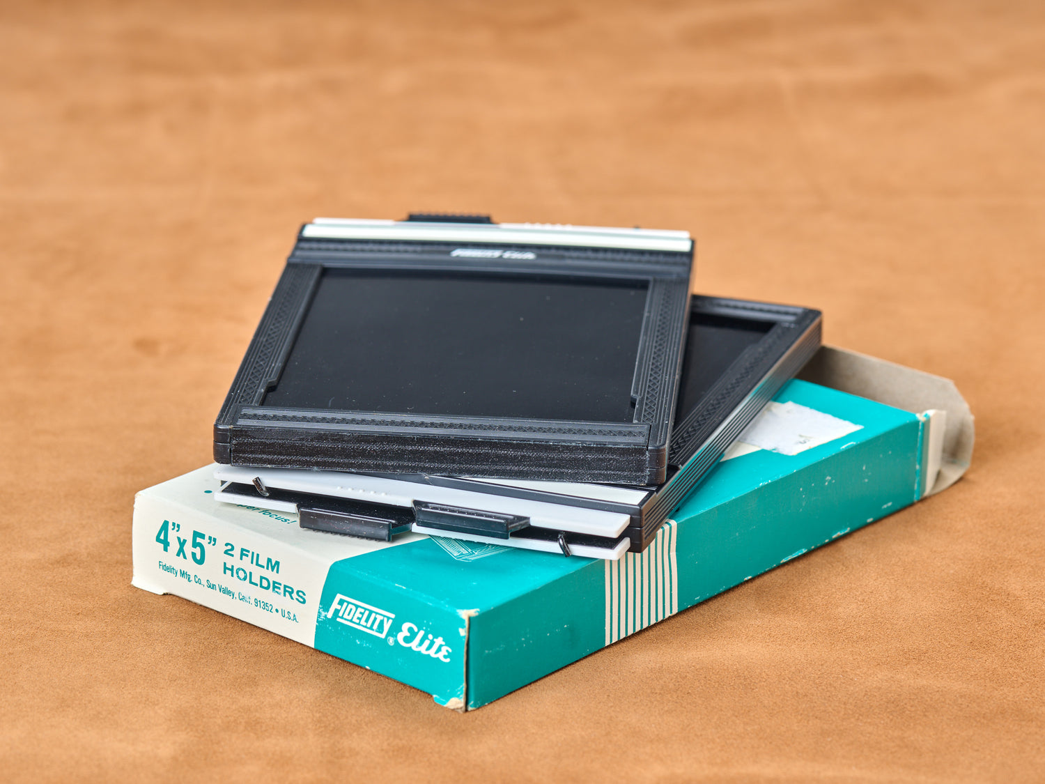 Black 4x5 film holder slips from teal box. White tabs beg to be pulled. Dark slide out, film exposed, hungry for light. Load. Lock. Fire twice.