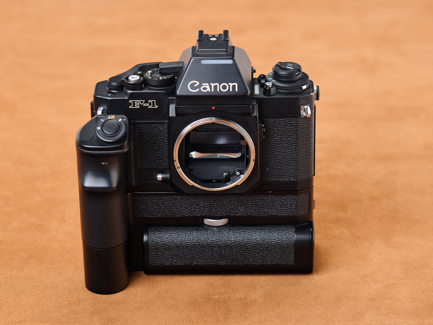 Canon new F-1 with Motor Drive, black body on suede. Vertical grip curves like a whispered promise. Shutter dares. Light glints. Ready to capture secrets.