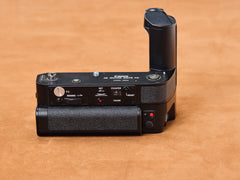 Canon new F-1 Motor Drive on suede, black body breathing heat. Rewind whirrs, counter ticks, red lights wink. Vertical grip rises, taut. Shhh. Come closer now.