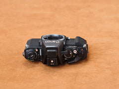 Canon F-1, black and hungry on suede. Shutter dial murmurs, film lever slides shhh. Silver winks in low light. Every curve hums, mechanism breathes. Slow tension.