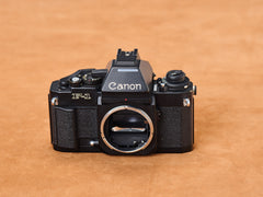 Canon F-1 from shadow, black lacquer gleaming. Chrome mirror box glints forbidden silver. FD mount yawns, hungry, exposed. Mirror slaps. You and machine, alone.