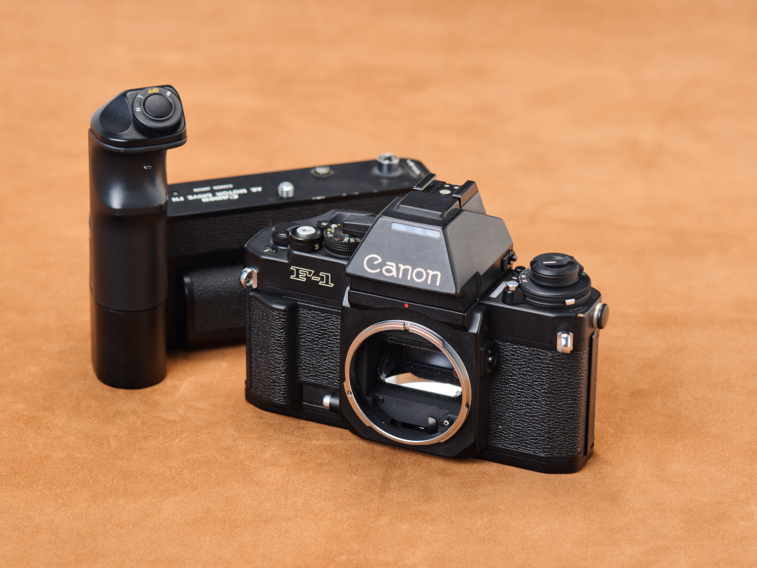 Canon new F-1, silver FD mount gleaming like a secret. Black motor drive detached, patient in shadow. Metal on tan suede sighs. Every curve whispers anticipation.