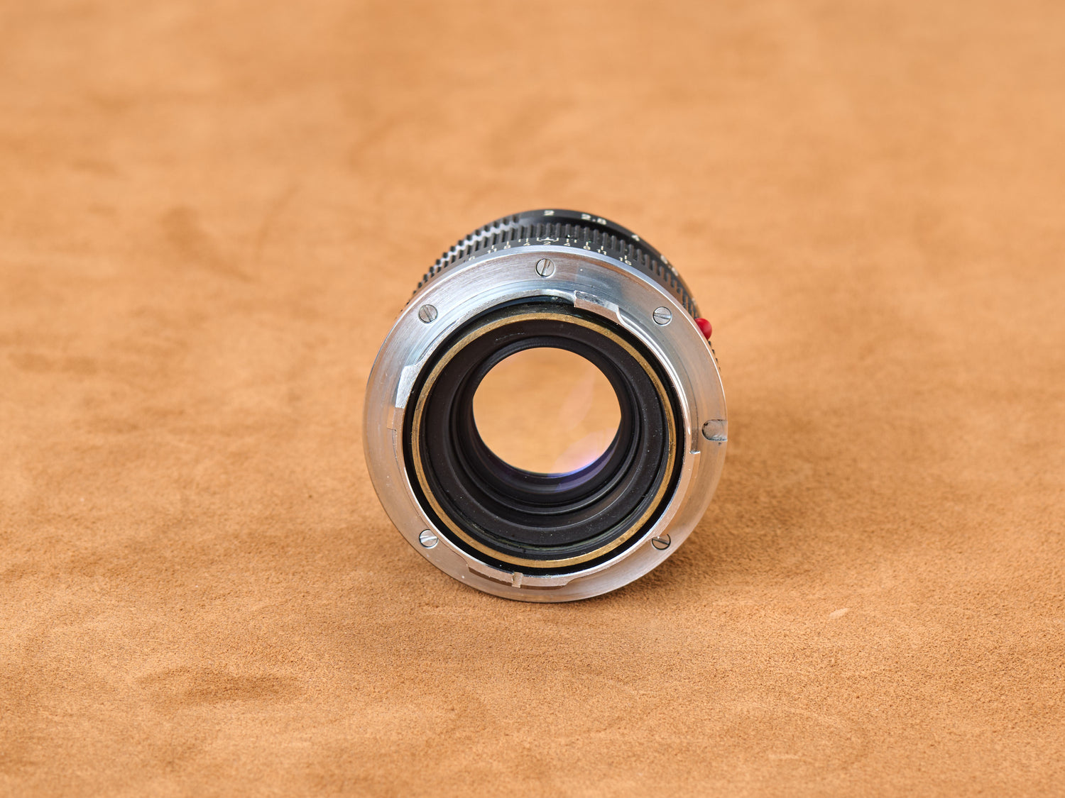 Click. The Leica Summicron 50mm f/2 exposed—chrome mount gleaming, brass coupling ring glowing gold beneath. Rear glass element waits, vulnerable, open. Red mounting dot burns on the barrel, a warning, a promise. Twist. The coupling mechanism spirals, precision machined to perfection. Tan suede beneath, warmth against cold metal. Studio light traces every thread, every curve. Touch the chrome. Feel the weight. Snap. Mount it. Lock it in. Let the rangefinder couple. Focus aligns. Perfect. Sharp. Inevitable.
