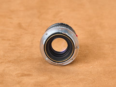 Click. The Leica Summicron 50mm f/2 exposed—chrome mount gleaming, brass coupling ring glowing gold beneath. Rear glass element waits, vulnerable, open. Red mounting dot burns on the barrel, a warning, a promise. Twist. The coupling mechanism spirals, precision machined to perfection. Tan suede beneath, warmth against cold metal. Studio light traces every thread, every curve. Touch the chrome. Feel the weight. Snap. Mount it. Lock it in. Let the rangefinder couple. Focus aligns. Perfect. Sharp. Inevitable.